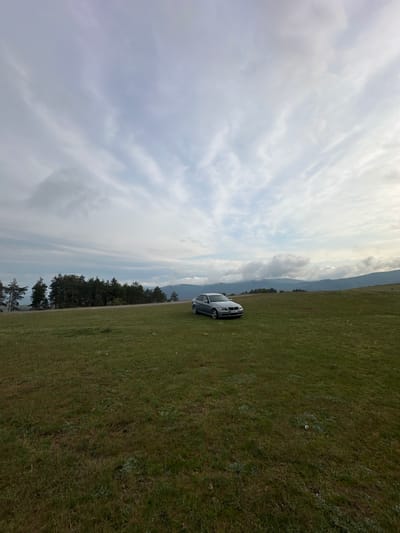 The image depicts a serene natural landscape, likely in Mirkovo, Bulgaria, under a cloudy sky. The foreground is dominated by a vast, green grassy field with some lighter patches of vegetation. A silver or light-colored sedan, appearing to be a BMW, is parked on the edge of this field, facing towards the distant mountains. Its license plate, "CA 6348 HB", confirms a Bulgarian registration.
Beyond the grassy expanse, a distinct line of dark green coniferous trees stretches across the midground. In the far distance, layers of rolling hills or mountains are visible under a sky filled with various cloud formations, ranging from thin, wispy clouds to more dense, overcast patches. The lighting suggests it is either late morning or afternoon on a partially cloudy day. There are no people visible, indicating a quiet and undisturbed natural setting. The scene evokes a sense of calm and openness, characteristic of a rural, mountainous or hilly region.