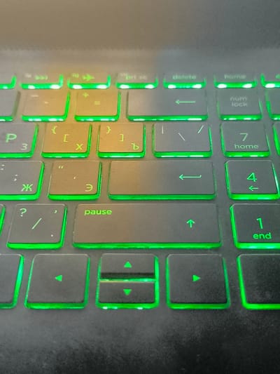 The image shows a close-up of a laptop keyboard with green backlighting. The keys visible include "P", "X", "Ж", "?", "/", "pause", "delete", "home", "num lock", "7", "4", "1", "end", and various symbols and Cyrillic characters. There are also arrow keys and media control keys visible. The keyboard is black, and the surrounding laptop casing is also black and appears to be textured. The lighting is dim, with the green backlight being the primary light source. The image is taken from a slightly elevated angle, looking down at the keyboard. There is a warm glow, likely from ambient light, in the upper left portion of the image. There is no indication of a specific location in the city of Makho, Georgia.