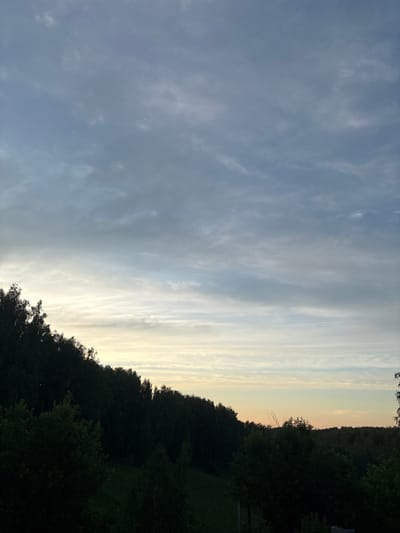 This is a vertical, eye-level, medium shot of a landscape. The image is dominated by a sky filled with thin, wispy clouds that stretch across the frame. The clouds are light gray and white, with some areas appearing brighter where the sunlight is breaking through. To the right of the sky, a dense line of dark green trees forms a silhouetted horizon. The trees are tall and lush, with a few taller ones jutting slightly above the rest. Beyond the trees, a faint, pale orange glow can be seen, suggesting the sun is low on the horizon. The overall mood is serene and peaceful. The time of day appears to be either sunrise or sunset. The location is identified as Maiskii, Russia. No people or objects are visible in the image, nor is there any visible text.