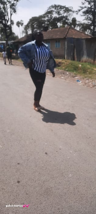 The image captures a sunlit street scene in Homa Bay, Kenya. In the mid-ground, a dark-skinned adult female is depicted in motion, walking leftward across a paved road. She is dressed in an unbuttoned light-blue denim jacket, a black and white vertically striped long-sleeved top, dark trousers (possibly jeans with a tear on the left thigh), and light-colored open-toed sandals. Her head is slightly turned towards the left of the frame. A prominent dark shadow of the subject is cast on the asphalt road surface to her right, indicating bright overhead sunlight.
In the blurred background, on the left side of the road, two additional individuals, one appearing to be an adult and the other a child, are walking away from the camera. To the right of the background figures, a single-story building with a reddish-brown tiled roof and light blue window frames is visible, situated next to a structure clad in corrugated metal sheeting. Various trees with dense foliage are scattered along the horizon, under a light-colored sky. The road is asphalt, with some minor litter visible along the right curb. Overlaid on the bottom left corner of the image is white text stating "police kombo" followed by two pink heart emojis.