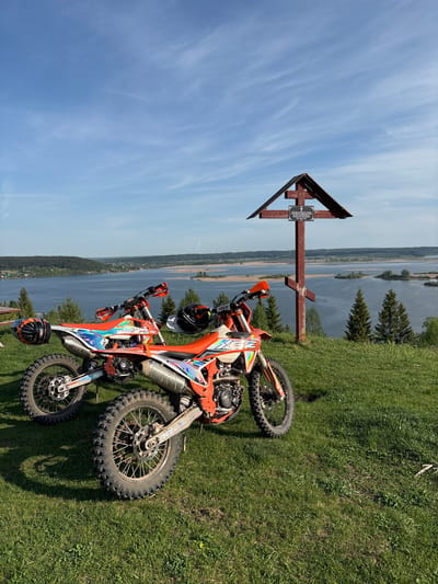 The image shows two orange dirt bikes lying on a grassy hillside next to a large wooden cross. The cross has a sign in Russian affixed to it. In the background is a body of water with forested areas along the shoreline. The location is likely the city of Serga, Russia, based on the language of the sign. The sky is blue with wispy clouds. Two black and orange helmets are placed on or near the bikes. The scene suggests a recreational or memorial site.