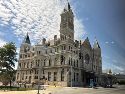 This is an outdoor, eye-level, wide-angle shot of Louisville Union Station in Louisville, Kentucky, USA, on a sunny day with scattered clouds. The grand, historic building, constructed from light-colored stone, dominates the frame. Its architecture features a tall clock tower with two clock faces visible, multiple spires, arched windows, and ornate detailing. A large circular stained-glass window is present on the facade of the main building, above a covered entrance area.
In the foreground, a street corner with asphalt and a raised curb is visible. A metal fence encloses a grassy area. Traffic lights are positioned at the intersection, with one showing a red light. A yellow fire hydrant stands near the sidewalk. A historical marker is placed on a small pedestal behind the fence. A large green tree with dense foliage is to the left of the building. The sky is blue with a pattern of fluffy white cumulus clouds. Reflections are visible on the surface from which the photograph was taken, suggesting it was shot through a window. A blue and white sign with the word "RAPID" is visible near the entrance to the station. A few bicycles and people can be seen near the station entrance and in the background.