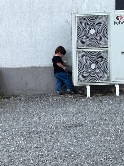 A young child, appearing to be a toddler, is standing next to an outdoor air conditioning unit in an outdoor urban environment. The child is wearing a black t-shirt with orange trim on the sleeves and collar, blue denim jeans, and brown shoes. The child is looking down and appears to be interacting with something near the base of the air conditioning unit, possibly holding a small object in their hands. The air conditioning unit is white and has two large circular grilles. The brand name "kobe" is visible on the unit. The ground is covered in gravel. The background consists of a textured white wall and a darker gray lower section of the wall. The scene is likely located in the city of Slanchevo, Bulgaria, given the context.