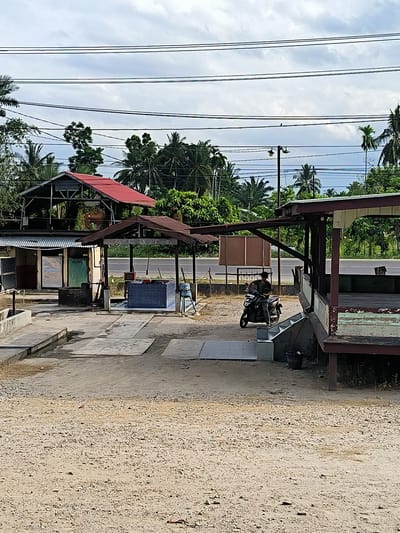 The scene shows an outdoor area in Blangbitra, Indonesia. A person is sitting on a motorbike next to a structure resembling a small shop or covered seating area, which is elevated. In the background, there is a series of open-air structures with corrugated metal and red roofing. A small building with doors is on the left. In the foreground, the ground is dirt and gravel. Above, utility lines stretch across the sky. Vegetation, including palm trees, is present in the midground and background.