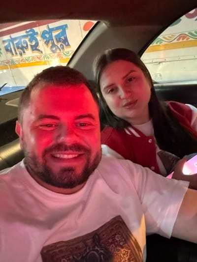 Two individuals are captured in a selfie-style photograph taken inside a vehicle. A male subject occupies the foreground, positioned centrally, smiling directly at the camera. He has a full beard, short dark hair, and is wearing a white t-shirt featuring a patterned design resembling a carpet or tapestry. His face and upper body are strongly illuminated by a red light source. Behind and to the right of the male, a female subject is partially visible, looking towards the camera with a neutral expression. She has long dark hair and is wearing a red and white jacket over a white garment. Her right hand is holding a smartphone, its screen emitting a faint light. The interior of the vehicle is dimly lit, with dark upholstery visible. Through the rear window, a larger white vehicle, likely a truck or bus, is clearly visible, displaying text in Bengali script: "বাক্স ই ফুল" (Bakso-i Phul), which translates to "Box is full." This external vehicle's presence and the Bengali script confirm the location as Bidhannagar, India. The image captures a momentary interaction between the two individuals within a car, adjacent to street traffic.
