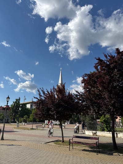 This image captures a bright, sunny daytime scene in an urban square or park within Becej Municipality, Serbia. The upper portion of the frame is dominated by a clear blue sky with scattered white cumulus clouds. Prominently in the mid-background, a white church steeple, topped with a cross, rises towards the sky. To its left, a modern building with multiple communication antennas on its roof is visible.

The ground is primarily paved with a pattern of light and reddish-brown rectangular pavers, interspersed with grassy areas. Several dark-colored wooden benches with black metal frames are positioned along the paved walkways and grassy sections. On the far left, a black ornate street lamp post, adorned with a hanging basket of pink and red flowers, stands.

Three trees with distinctive dark red/maroon foliage are scattered across the mid-ground, two larger ones framing the right side of the image and a smaller one closer to the center. These trees and the street lamp cast long, distinct shadows on the paved ground, indicative of strong sunlight.

Four individuals are present in the scene, all moving from left to right. In the left-midground, a person, likely male, rides a bicycle, wearing a light-colored short-sleeved shirt and light pants, with a white bag hanging from the handlebars. Further to the right and slightly behind, a group of three people is seen: one person pushes a baby stroller, another person walks alongside, seemingly talking on a mobile phone, and a third person on a bicycle follows behind them. In the far background, large white letters on a low wall are partially visible, suggesting a landmark or city name.