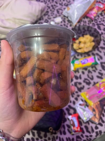 A hand holds a clear plastic cup filled with small, irregular brown pieces that appear to be snacks. The cup has a clear plastic lid with a small red tab. The background is a blurry leopard-print fabric. Several colorful snack bags and a small dark purse are also visible in the background, suggesting a casual setting, possibly at home or while relaxing. The lighting is artificial, casting reflections on the plastic cup and creating purple light streaks. There is no visible text in the image. The time of day and weather are indeterminate.