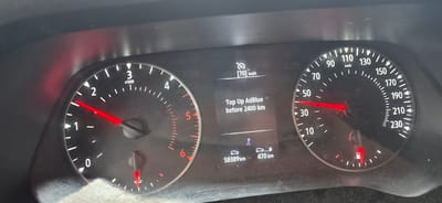 The image shows a close-up of a car's dashboard, specifically the instrument cluster and a digital display. The primary objects are the tachometer on the left, the speedometer on the right, and a central digital screen.

The tachometer has red needles and markings from 0 to 6, with "x1000" indicating the engine RPM. The needle is positioned just above the '1', suggesting the engine is idling or running at a low RPM. The speedometer has markings from 0 to 230 km/h, with the needle indicating a speed of around 30 km/h. A fuel gauge is visible at the bottom right of the speedometer, showing a nearly full tank.

The central digital display shows a speed of [70] km/h, and a warning message: "Top Up AdBlue before 2400 km". Below this, there are icons representing a car and a fuel pump, with corresponding mileage figures: "58389 km" and "470 km". This indicates the total mileage of the car and the estimated remaining distance based on the current fuel level.

The scene is indoors, likely within the cabin of a vehicle, as indicated by the dashboard elements. The lighting appears to be from daytime, possibly overcast given the lack of strong shadows, suggesting it is not a bright sunny day. There are no people visible in the image. The dashboard itself shows some dust and smudges, typical of a used vehicle. The location is identified as Aarhus, Denmark. The overall mood is functional and informative, conveying vehicle status and maintenance alerts.