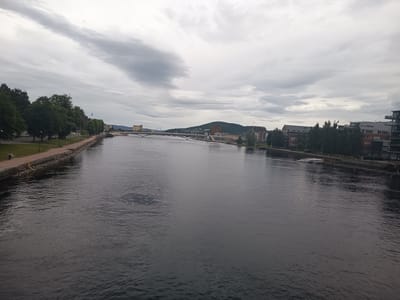 The image depicts a wide river in Drammen, Norway, under a cloudy sky. The river dominates the foreground, its dark, rippling surface reflecting the overcast sky. On the left bank, a paved walkway runs alongside the water, bordered by a stone embankment and lush green trees. A few lampposts line the walkway. Further along the left bank, a small, red vehicle is visible.
The middle ground is characterized by a long, arched bridge spanning the river. Beyond the bridge, a cityscape unfolds. On the right bank, modern apartment buildings with glass balconies and brick facades are visible, interspersed with trees. A white boat is docked along the right bank. In the distance, a hill rises, topped with a building and a visible antenna. The overall atmosphere is tranquil, suggesting a late afternoon or early evening on a cool, overcast day. There is no visible text in the image.