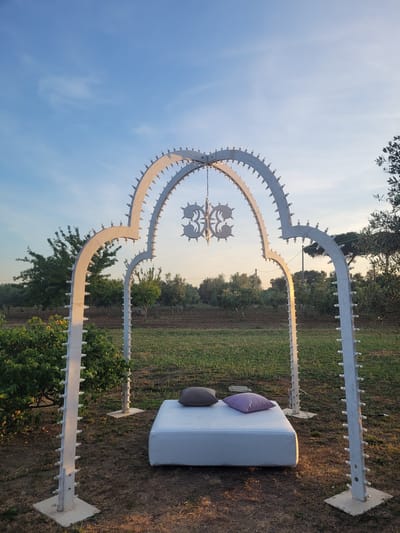 This image captures an outdoor scene in Sogliano Cavour, Italy, likely during the golden hour of late afternoon or early evening, evidenced by the warm, golden light illuminating parts of the scene and the bright, clear sky.

The central subject is a large, ornate, light-colored metal archway standing proudly in a grassy, rural setting. The archway features two main curved supports that meet at the top to form a symmetrical, stylized heart or clover-leaf shape. Numerous small, clear light bulbs are affixed along the edges of this structure, suggesting it's designed to be illuminated, possibly as a "luminarie" or festive decoration. In the very center of the arch, where the curves converge, there is an intricate, smaller metal embellishment, also equipped with light bulbs, resembling a floral or geometric motif. The entire structure is anchored to the ground by two substantial, light-colored concrete or stone bases.

To the right of the archway, a white, rectangular outdoor bench or sofa is positioned, offering a place for relaxation or viewing. Two cushions adorn the bench: one purple and one dark grey or brown. The ground around the arch and bench is a mix of dry grass and darker earth.

In the background, beyond the decorative archway, lies an expansive rural landscape. Rows of green trees, likely olive groves, stretch into the distance, interspersed with patches of cultivated land. A utility pole can be seen in the middle ground. The overall impression is one of a tranquil, prepared outdoor space, possibly for an event, a garden feature, or a photo spot, basking in the soft glow of the setting sun. No people are visible, and there is no visible text.