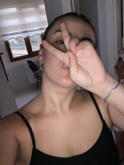 In an indoor setting, a young woman, with her face partially obscured by her right hand forming a peace sign, looks towards the camera. She is wearing a black tank top and a silver bracelet with a blue evil eye charm. Behind her, a window with horizontal blinds reveals a view of a street with parked cars and trees. A radiator is visible beneath the window. In the background, a person with a towel wrapped around their head is partially visible. The overall lighting is dim. The scene appears to be located in Yunus Emre, Turkey.
