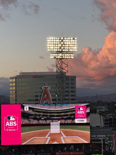 This image captures an atmospheric scene at a baseball stadium during dusk or twilight. The sky transitions from a soft blue-grey to warm pink and orange hues with scattered clouds, indicating the setting sun.

In the foreground, a large, brightly lit video scoreboard dominates the lower half of the frame. It displays a graphic of a baseball field from behind home plate, with a transparent strike zone box and a small baseball visible. The scoreboard features prominent branding: on the left, a magenta panel shows "ABS" with a baseball player logo, "Powered by T Mobile," and a small "A" logo. A similar magenta panel on the right displays "ABS," "T Mobile," and "• STRIKE," also with a small "A" logo.

Above the scoreboard, a tall light tower with numerous rows of brilliant white stadium lights illuminates the scene. Behind the light tower and the scoreboard is a large, modern building with a prominent red "A" logo with wings, characteristic of the Los Angeles Angels baseball team. The building itself has "Impact" visible as signage.

Below the scoreboard, smaller text advertisements are visible on a barrier, reading "NITTO TIRE.COM" (repeated) and "SALINAS, INC." (repeated). Further to the right, a "TEAM STORE" sign can be seen. In the distance, rolling hills or mountains are silhouetted against the fading light. The overall scene suggests the beginning or end of a baseball game, set against a picturesque sunset at Angel Stadium in Anaheim, United States.