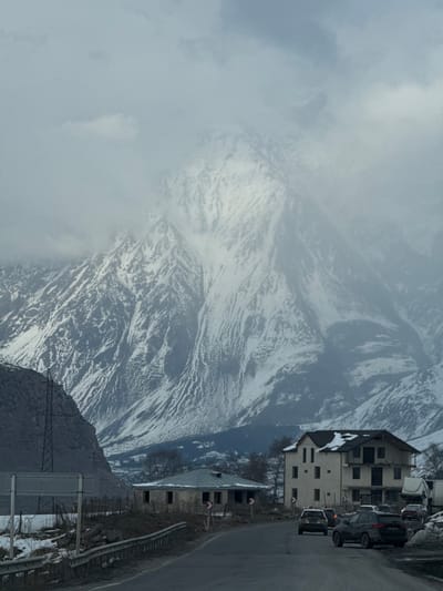 This image depicts a mountainous, wintery scene in Garbani, Georgia, featuring a paved road and several buildings under an overcast sky.
In the background, a massive snow-covered mountain range dominates the upper portion, with rugged peaks and slopes heavily layered in white snow. Dense, grey clouds partially obscure the mountain tops and hover over the upper ridges, limiting visibility.
In the midground, a dark asphalt road curves from the lower-left foreground toward the right. Adjacent to the road on the right, a multi-story building with a light-colored facade and a dark, gabled roof stands, exhibiting some snow accumulation on its roof. Multiple windows are visible, and construction elements or partial finishes are apparent on its exterior. To its left and further down the road, a single-story building with a dark roof and light walls, appearing to be unfinished, is situated. Bare deciduous trees are sparsely scattered around the buildings and in the valley behind them.
In the foreground on the left, a grey metal guardrail runs alongside the road, partially covered with dirty snow and bare earth. A large metal electricity transmission tower is positioned on the far left, against a darker, rocky lower mountain slope. Brown arrow signs indicating a right curve are visible near the single-story building. Several vehicles are present on and alongside the road; a dark SUV is prominently visible in the lower-right foreground, facing away from the viewer, with other cars and a white truck or van parked further up near the multi-story building.
The environment is cold and overcast, with visible snow on the ground and mountains, consistent with winter conditions. No individuals are discernible.