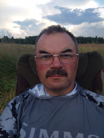 A middle-aged man with a mustache, wearing glasses and a gray camouflage shirt, is sitting in a chair outdoors. The background shows a field with tall grass, trees, and a cloudy sky. He is looking directly at the camera. The image appears to have been taken in a rural area, possibly in the city of Garanski Rural Council, Belarus.