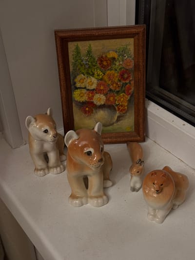 A static image displays a collection of decorative items positioned on a white, textured surface, likely a window sill or shelf, within an unspecified building in Zelenogradskiy, Russia. To the left, a light-colored wall is visible. To the right, a dark window frame and glass panel are present.

The scene features four ceramic animal figurines and one framed painting. From left to right:
1.  A ceramic figurine, resembling a tan and cream-colored lion cub or fawn, stands facing slightly right.
2.  A second, similar ceramic figurine, also tan and cream-colored, stands slightly in front and to the right of the first, facing forward and slightly left.
3.  Behind and between these figurines, a rectangular painting in a dark wooden frame stands vertically. The painting depicts a still life of a bouquet of colorful flowers, predominantly orange, red, and yellow, with green foliage, arranged in a grey vase against a light brown background.
4.  To the right of the central lion cub figurine, a smaller ceramic figurine resembling a grey and tan squirrel stands upright.
5.  Furthest to the right, a fourth ceramic figurine, resembling a tan and cream-colored lion cub or small dog, lies or sits facing left. This figurine appears to be a salt or pepper shaker, indicated by subtle features.