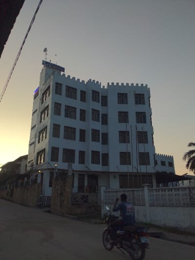 This image depicts a street scene in Bagamoyo, Tanzania, likely during the late afternoon or early evening. The sky, which dominates the upper left, transitions from a light grey-blue to a warm, golden hue near the horizon, indicative of sunset.
The central subject is a tall, multi-story white building characterized by a distinctive castellated parapet along its roofline, giving it a castle-like aesthetic. Its facade is punctuated by numerous rectangular windows. On the upper left side of the building, a blue sign is visible, with a portion of the text appearing to read "BAGAMOYO," a clear location cue. Another smaller blue sign is located further down the building's side.
In the foreground and to the right, a paved street runs alongside the building. A person is seated on a motorcycle on the street, facing away from the camera. A decorative, white patterned wall or fence separates the building's property from the street. The top right corner shows a glimpse of palm tree fronds. The scene appears calm and everyday, with the warm light casting a soft glow over the surroundings.