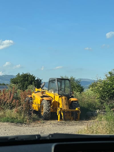 A yellow JCB telehandler is parked on a dirt road, partially obscured by overgrown vegetation. The telehandler is facing forward, with its boom retracted. The vehicle is positioned in an outdoor setting with a blue sky and scattered clouds overhead. In the background, there are rolling hills covered in trees, and some solar panels are visible to the left. The foreground shows the road surface and the dashboard of a vehicle from which the photo appears to be taken. The location is identified as Gorni Bogrov, Bulgaria. There are no people visible in the image.