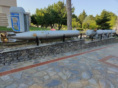In Litochoro, Greece, a public display features a torpedo and a series of mines. The torpedo, positioned on the left, is a cylindrical, light-gray object with a prominent brass propeller at its rear. It is labeled with "DANGER DO NOT TOUCH" and "KINAYNOΣ" in Greek. Behind the torpedo, a light-gray structure resembling a modified telephone booth with blue accents and the "OTE" logo is visible. To the right of the torpedo, several cylindrical, gray mines are arranged in a line, connected by chains. These mines appear to be on display on a low, stone-faced wall. The ground in the foreground is paved with irregular, flat stones, with a red brick border delineating a path. In the background, lush green trees and a clear blue sky suggest a pleasant outdoor setting. A large tree trunk is prominent in the middle ground.
