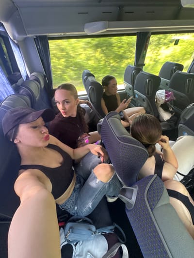 The image captures the interior of a moving passenger coach bus during daylight hours. The scene is located in the city of Nikolaevo, Bulgaria. Four young adult women are visible, occupying multiple seats and the aisle space.
In the immediate foreground, a woman wearing a black baseball cap, black tank top, and ripped blue jeans is taking a selfie. She has dark hair, prominent red lipstick, and is making a puckered lip expression towards the camera. Her right arm is extended, holding the camera, and her left arm rests on her bent knee. A light blue shoulder bag with a gold chain strap rests on the floor near her left leg.
To her right and slightly behind, a second woman, wearing a dark reddish-brown long-sleeved top and ripped blue jeans, looks directly at the camera with a slight smirk. Her dark hair is pulled back, and she holds a clear plastic bottle with a pink cap in her right hand.
Further back, seated by a window, a third woman with dark hair pulled back in a bun or ponytail is engrossed in her smartphone, looking down at the screen. She wears a black sleeveless top and has bare legs crossed at the ankles.
Partially visible in the background, behind a seat, a fourth woman with dark hair in a low ponytail or bun is seen with her hand near her ear, holding a dark pen-like object. She also wears a black top.
The bus interior features dark blue patterned seating with grey trim, grey overhead storage compartments, and large windows revealing blurred green foliage, indicative of travel through a vegetated area. Blue patterned curtains are partly drawn on the left side of the bus. A white cloth or plastic bag is placed on a seat in the far right background.