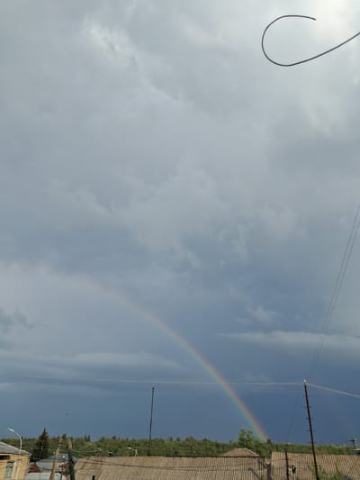 The image depicts a partially visible rainbow arching across a cloudy sky above a residential area in Gavar, Armenia. The sky is a mix of light gray and darker, stormy clouds. A curved wire, possibly a power line, is visible in the upper right corner of the frame. Below the sky, a line of trees and greenery forms a backdrop for a row of houses with corrugated roofs. Electrical poles and wires are scattered throughout the scene, indicating a developed urban or semi-urban environment. A bird is faintly visible in the darker sky in the middle left of the image. The lighting suggests it is daytime, likely after or during a rain shower.
