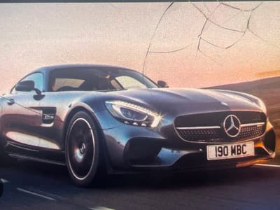 The image shows a dark grey Mercedes-Benz sports car with the license plate "190 MBC". The car is in motion, likely on a road, with a blurred background suggesting speed. The sky is a warm gradient of orange and pink, indicating either sunrise or sunset. There are visible cracks on the screen or lens used to capture the image, overlaying the scene. No people are visible in the image. The location in Paczkowo, Poland is not identifiable from the visual information provided.