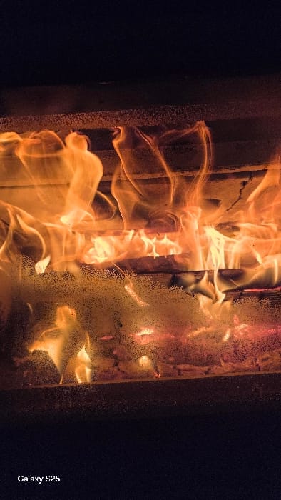 The image displays an active fire contained within a structure, identified as being in Ottonville, France. Multiple wooden logs are observed burning, generating prominent orange and yellow flames that rise vertically. Glowing red embers are visible beneath the active flames. A semi-transparent surface, likely a glass barrier, is positioned in the foreground and shows numerous small, diffuse light particles scattered across its expanse, partially obscuring the view of the fire. The area above and below the firebox is dark, consistent with a low-light environment. No individuals, actions, or interactions are present in the scene. A "Galaxy S25" watermark is located in the lower-left corner.