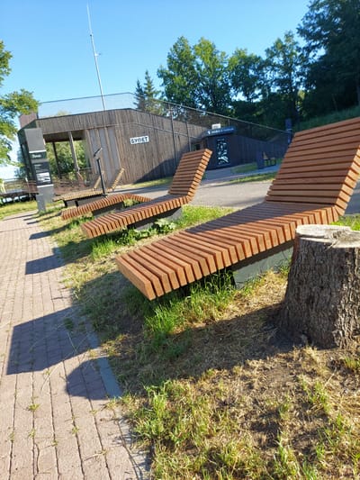 In the city of Razvetye, Russia, on a clear, sunny day, a park scene unfolds with a focus on modern outdoor seating. The foreground is dominated by a large, rough tree stump on the right, with its bark clearly textured. Adjacent to it, a paved walkway made of interlocking bricks runs diagonally from the bottom left, casting a distinct shadow. To the left of the walkway, dry grass and sparse green shoots cover the ground, indicative of a well-trodden area.
Several contemporary wooden lounge chairs, designed with horizontal slats, are arranged in a row, facing away from the camera and towards a body of water or a scenic view, which is partially visible in the background. These chairs are set on dark bases, contrasting with the natural wood color and the surrounding greenery.
Behind the seating area, a modern wooden structure with a slatted facade and a metal railing stands on an elevated platform. This building appears to be a pavilion or a small cafe, as indicated by signage with Cyrillic text. One sign reads "БУФЕТ" (bufet), meaning "buffet" or "canteen," and another displays "кофе с собой" (kofe s soboy), translating to "coffee to go." The building is situated on a grassy incline, with mature trees providing a lush backdrop against the bright blue sky. The overall scene suggests a public leisure area designed for relaxation and enjoyment of the outdoors.