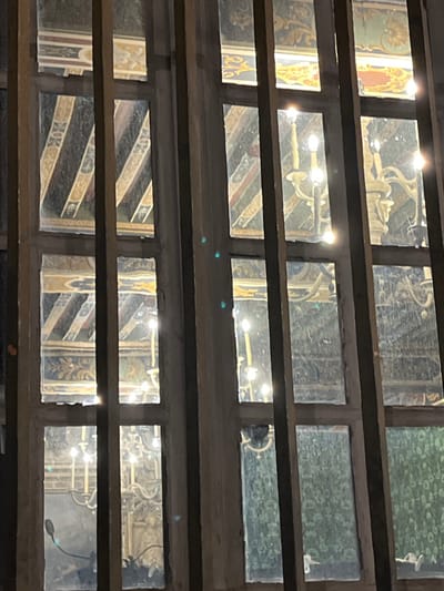 A multi-paned window offers a glimpse into a space adorned with ornate decorations. Chandeliers hang from the ceiling, reflecting light throughout the room. The walls feature intricate designs and patterns, suggesting a historical or religious setting. The view is partially obscured by the window frame, creating a layered perspective.