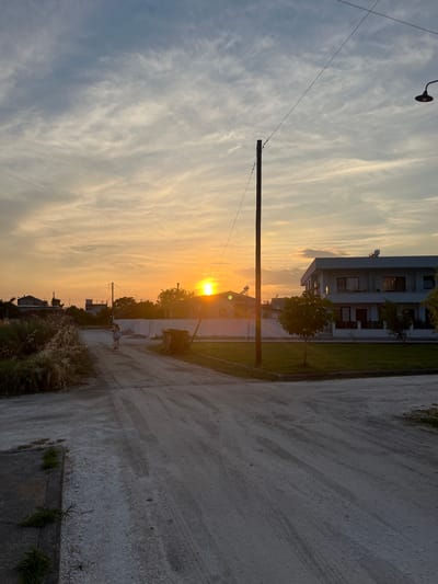 This is a vertical, eye-level, outdoor photograph taken at sunset in Chaidefto, Greece. The scene depicts a residential area with a dirt road running through it. A modern, white, multi-story building with balconies is visible on the left side of the road, with a small tree planted in front of it. The road itself is textured with tire marks and has a concrete border with some grass growing on it.

The sun is low in the sky, casting a warm, golden glow across the scene, illuminating the clouds which are wispy and stretch across the sky. A utility pole with electrical wires is prominent in the foreground, with a light fixture attached to it. In the distance, other houses and trees can be seen, suggesting a suburban or rural setting. A lone figure, a young girl in a light-colored dress, is walking on the dirt road, heading away from the camera. The overall mood is peaceful and serene, characteristic of a quiet evening. The time of day is clearly sunset due to the quality of light and the position of the sun. There is no visible text in the image.