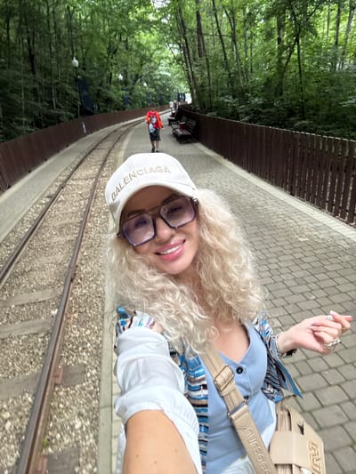 In the city of Guamka, Russia, a woman with curly blonde hair, wearing a white cap with "BALENCIAGA" written on it, glasses, a blue shirt, and a colorful patterned jacket, poses for a selfie. She is standing on a paved platform next to a railway track. In the background, trees form a dense forest, and a wooden fence runs along the platform. A few people, including a child in a red shirt, are visible in the distance. The scene appears to be a tourist location, possibly a park or a scenic route.
