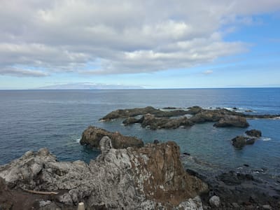 The image depicts a rugged coastal landscape overlooking the Atlantic Ocean. In the foreground, large, weathered rocks of varying textures and colors dominate the scene, with some exhibiting a brownish, iron-rich hue and others a lighter grey. The rocks are partially covered in dark soil and sparse vegetation.

Beyond the rocky shoreline, the deep blue ocean stretches towards the horizon. Several dark, jagged rock formations emerge from the water, creating a dramatic contrast with the calm sea. Small waves lap against these submerged rocks, producing white foam.

In the distance, a long, low-lying island or landmass is visible on the horizon, its outline softened by a hazy atmosphere. The sky is filled with a mix of grey and white clouds, with patches of clear blue sky peeking through, suggesting a partly cloudy day. The lighting is soft, indicating it might be overcast or late in the day.

The scene is located in Puerto De Santiago, Spain, a coastal town known for its volcanic landscapes and ocean views. The overall impression is one of natural beauty and raw, untamed coastal scenery. There are no people or animals visible in the image.