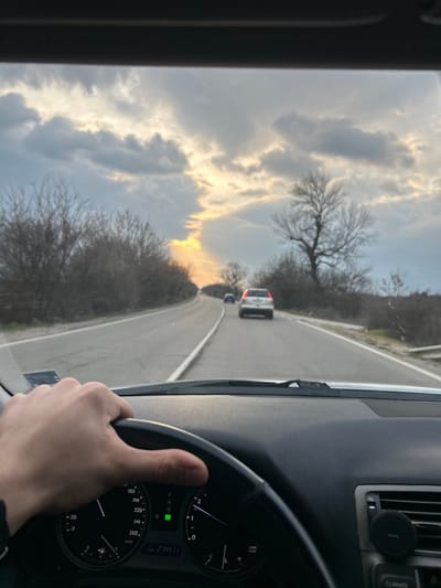 The image presents a first-person view from inside a vehicle, likely from the driver's position, traveling on a road in Stolnik, Bulgaria. A pale-skinned left hand is visible on the black steering wheel, positioned at approximately the 9 o'clock mark. The vehicle's dark grey dashboard is in the foreground, displaying a speedometer indicating approximately 60 km/h and an odometer reading 274411. The illuminated 'D' confirms the vehicle is in Drive. A circular black phone mount is affixed to an air vent on the passenger side of the dashboard.
The two-lane asphalt road ahead curves gently to the left, marked by a solid white center line and a broken white line on the right shoulder. Two vehicles are visible in the distance, traveling in the same direction: a silver-colored SUV or station wagon is closer, followed by a darker blue sedan. Both sides of the road are lined with dense, bare deciduous trees and shrubs, consistent with late autumn or winter foliage. A prominent, large bare tree stands on the right side of the road in the midground.
The sky is predominantly overcast with grey clouds. A distinct, narrow vertical shaft of intense yellow-orange light breaks through the clouds in the center-left, indicating the low position of the sun, suggestive of either sunrise or sunset.