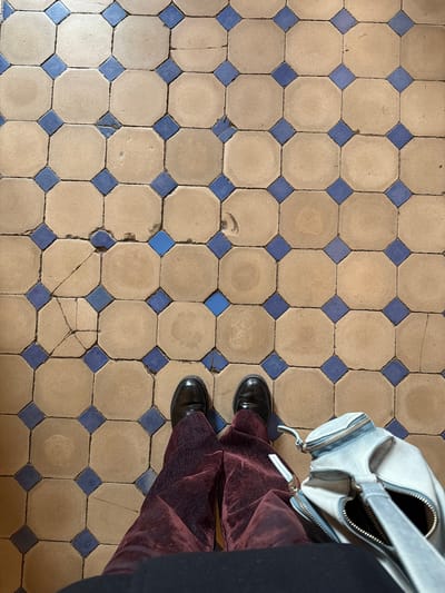 The image shows a tiled floor, likely indoors. The tiles are arranged in a grid pattern, with octagonal beige tiles surrounded by smaller blue square tiles. Some tiles show cracks and signs of wear. Two black shoes are visible near the top right of the image, suggesting a person is standing on the tiles. Part of a light-colored handbag is also visible near the shoes. The location is likely La Plata, Argentina, based on visual characteristics of the tiles and architecture.