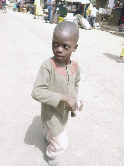 A young Black male child, approximately four to seven years old, stands on a light-colored, unpaved surface within a market area in Kadoma, Zimbabwe. He has a close-cropped haircut and wears an olive-green long-sleeved tunic with reddish-brown horizontal stripe detailing on the chest placket, light-colored baggy trousers, and dark sandals. The child is looking towards the left of the frame with a slightly open mouth. His right hand grasps a crumpled, light-colored object, possibly paper or cloth. The background features a blurred, sunlit outdoor market scene with multiple adult pedestrians, various market stalls, sacks of goods, and provisional structures providing shade. Shadows from figures and objects are visible on the ground, indicating bright ambient light.