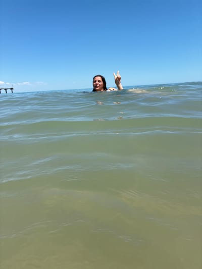 A woman is in the water at Shkorpilovtsi beach, Bulgaria, with her head and shoulders visible above the surface. She is smiling and holding up two fingers in a peace sign. The sky is clear and blue, and the water is calm with small waves. In the background, a pier can be seen.