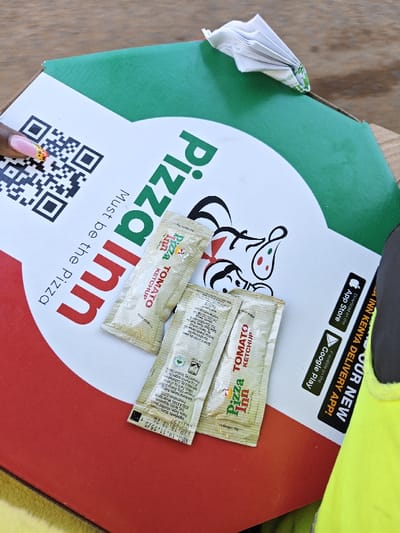 The image displays a close-up, top-down view of a Pizza Inn pizza box, indicating a meal either just delivered or about to be consumed. The pizza box is predominantly white in the center, with a large green arc at the top and a red arc at the bottom, typical of the brand's colors.

Prominently featured on the white section of the box is the "Pizza Inn" logo in green and red lettering, oriented vertically. Below the logo, the slogan "Must be the Pizza" is written in smaller, black text. To the left, a large QR code is visible, partially obscured by a person's hand. This hand has well-maintained fingernails painted with an orange and yellow, possibly cheetah print, design.

Resting on the center of the pizza box are three individual sachets of "Pizza Inn TOMATO KETCHUP". These sachets are light tan or off-white and feature the Pizza Inn logo along with the product name. Small print on the sachets indicates "Weight: 15g". On one of the sachets, an expiration date "EXP. 18.08.26" and a manufacturing date "MFG. 18.11.25/3" are clearly legible.

In the top right corner of the box, a crumpled white paper napkin is visible, tucked into the seam of the box. Towards the right side of the box, black text advertises the "OUR NEW Pizza INN KENYA DELIVERY APP!", with logos for "Download on the App Store" and "Available on the Google Play".

The background suggests a wooden surface, likely a table, where the pizza box is placed. A bright yellow item is partially visible on the far right, next to the pizza box. The overall impression is that of a casual dining or take-out experience.