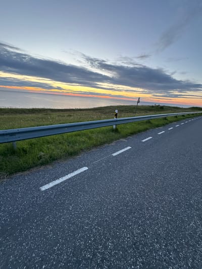 The image depicts a paved road running alongside a guardrail and a grassy embankment. The road has white dashed lines marking its lanes. Beyond the embankment is a body of water, likely the Baltic Sea, with a colorful sunset visible on the horizon. The sky is a gradient of blue and orange, with scattered clouds. A single traffic sign is visible on a pole in the distance. The scene appears to be located in Paldiski, Estonia, given the coastal setting and the typical road infrastructure. There are no people or animals visible in the image. The time of day is either dawn or dusk, as indicated by the sunset. The overall atmosphere is tranquil and scenic.