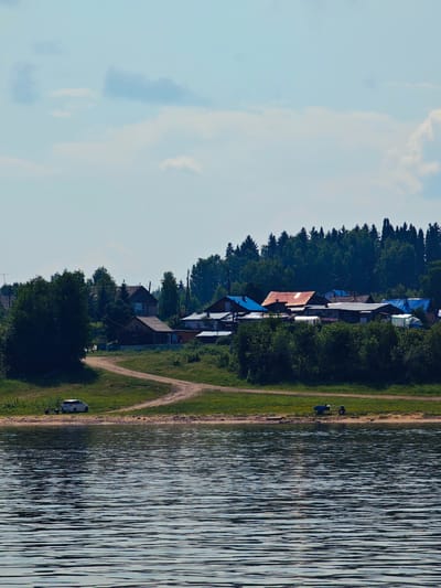 The image shows a rural village with several houses, some with blue and red roofs, nestled against a dense forest. A dirt road winds through a grassy area leading down to a body of water. A white car is parked near the water's edge, and a small blue trailer is also visible on the shore. The water in the foreground has ripples, suggesting a gentle breeze. The sky is partly cloudy. The scene is consistent with the location of Усть-Кемь, Russia, a village situated on the bank of a river.