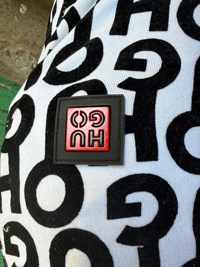A close-up, overhead shot captures a black patch with red lettering on a white t-shirt. The t-shirt has a bold, repeating black pattern of letters. The patch, which is square with rounded corners, displays the red letters "HU GO" arranged vertically, with "HU" above "GO". The scene is devoid of any specific location markers within Peshtera, Bulgaria, and the image focuses solely on the garment.