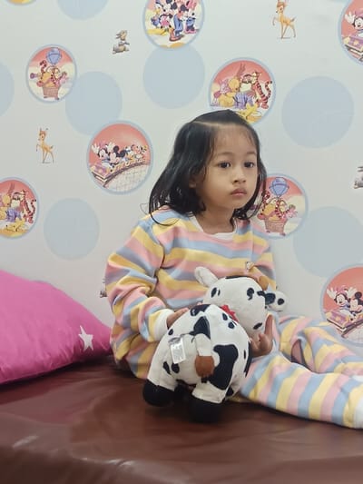 A young Asian female child, approximately 3-6 years old, is seated indoors on a brown cushioned surface in Karangtengah, Indonesia. She has dark, shoulder-length hair and wears pastel-striped pajamas featuring horizontal bands of light blue, yellow, pink, and white. The child holds two plush cow toys, both white with black spots; one is presented upright, showing its face, while the other is inverted, displaying its posterior, a brown tail, and a red ribbon on its head, along with a visible tag. To the child's left, a pink pillow with a white star design is partially visible. The background wall is white or light blue, decorated with circular decals depicting various Disney characters, including Mickey Mouse, Minnie Mouse, Winnie the Pooh, and Tigger, alongside individual character cutouts such as Bambi and Thumper. The child gazes forward with a neutral expression.