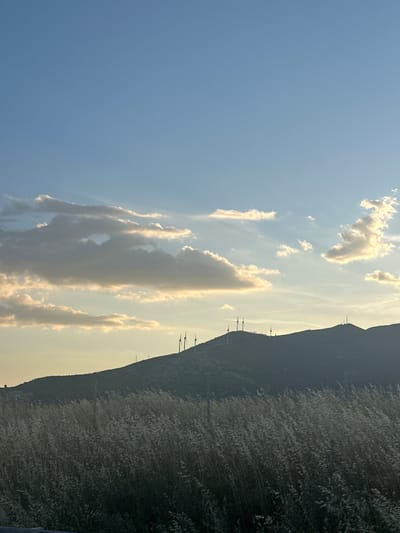 The image shows a landscape with a mountain range and a cloudy sky. The mountain is covered in dense vegetation. Silhouetted wind turbines are visible along the mountain ridge. The sky is blue with white clouds. The picture was taken in Manisa, Turkey.
