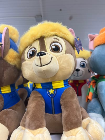 The image shows a close-up, vertically oriented, of several plush toys. The most prominent toy in the foreground is a brown and yellow dog wearing a blue and yellow uniform with a yellow star emblem. It has large, round eyes and a fuzzy yellow trim around its head. Other plush toys are visible behind it, including a white dog with blue eyes and a dog with an orange and blue scarf. The toys are stacked together, and the background appears to be a wall with horizontal lines, possibly a shelf or display unit. There is a small tag attached to the front of the main toy, with some text and graphics visible, but it is not legible. The lighting suggests an indoor setting. The image does not depict any people or specific events. The context of Ivanivka, Ukraine, does not provide further visual cues within the image itself.