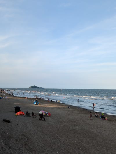 The image depicts a crowded beach scene in Hoằng Tiến Commune, Vietnam. The sea is calm with gentle waves breaking on the shore. Numerous people are visible in the water, swimming, wading, and enjoying the beach. On the sand, individuals and small groups are scattered, some sitting on chairs, others playing with beach toys. Several inflatable rings and a couple of colorful beach balls are present. In the foreground, there are stacks of black tires, orange life vests, and empty plastic chairs. Further down the beach, a person wearing a conical hat sits on a chair. In the distance, a small, verdant island with trees rises from the sea. The sky is a clear, pale blue with a few wispy clouds. The overall impression is a lively and popular beach destination.