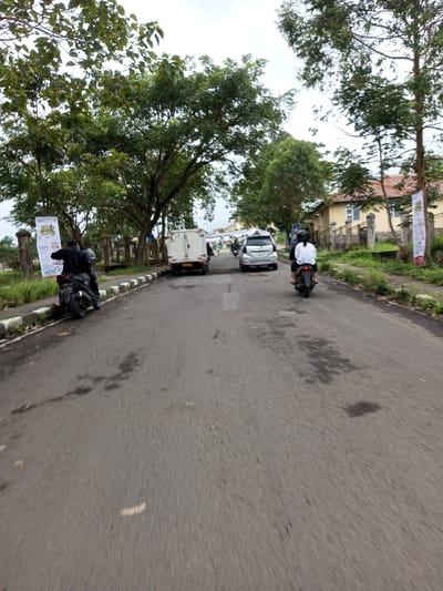 The image displays a two-lane asphalt road in Melakudikadu, India, under an overcast sky. The road surface shows areas of wear and dark stains. White-painted curbs line both sides of the road, separating it from grassy verges populated by numerous tall, green trees.
On the left side of the road, a person wearing a dark jacket and helmet is seated on a black scooter, stopped or parked near the curb, facing away from the camera. Immediately behind this scooter, a large vertical banner with colorful text and graphics is affixed to a pole. Further down the left side, a white box-style utility van is positioned, followed by a silver SUV, both oriented away from the camera.
On the right side of the road, a person in a white shirt and dark helmet is riding a black scooter, also moving away from the camera. In the background on the right, a light-colored building with a terracotta-tiled roof is visible behind a stone fence and additional trees. Another vertical banner is partially visible near the building on the right. The overall scene indicates active road use within a vegetated environment.