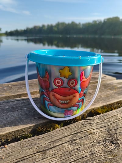 A close-up, eye-level shot focuses on a small, blue-rimmed plastic bucket with a cartoon crab design, resting on weathered wooden planks. The bucket's interior reveals a few grains of sand. The planks, weathered and gray, are partially covered by patches of green moss in the gaps between them. In the background, a calm body of water stretches towards a treeline under a clear blue sky with some wispy clouds. The scene is illuminated by natural sunlight, casting shadows on the wooden surface. The location is the shore of a lake in Brocēni, Latvia.