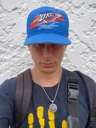 A man wears a blue Oklahoma City Thunder baseball cap, a black t-shirt with yellow designs, and a silver necklace with a circular pendant. He is also wearing a black backpack. The background is a white textured wall. The location, San José de Barlovento, Venezuela, is not discernible from the image.