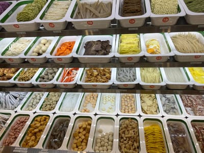 The image shows a display of various ingredients for hot pot, likely in a supermarket or restaurant. The ingredients are organized in white rectangular containers with green borders, each filled with a specific food item. These include different types of noodles (ramen, udon, glass noodles), vegetables (spinach, carrots), proteins (beef strips, chicken gyoza, meatballs, shrimp, squid, sausages, fish balls), and other items like quail eggs and tofu. Some containers have labels in German, such as "Ramen spinat," "Udonnudeln," and "Rinderstreifen." The display is tiered, with multiple shelves visible, suggesting a large selection. The lighting appears to be artificial, typical of an indoor setting. The overall scene is clean and organized, with the food items presented in an appealing manner for customers to choose from. The specific location in Bottrop, Germany, is not directly evident from the visual information within the image.