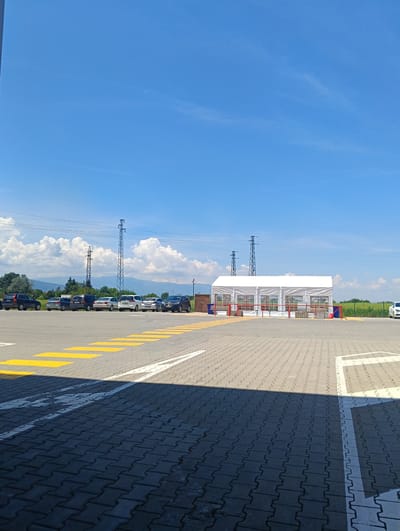 The image depicts a large outdoor event tent set up in a parking lot in Elin Pelin, Bulgaria. The tent is white and appears to be made of fabric with clear plastic windows. It is situated on a paved surface, with some red and blue barriers at its entrance. In the background, several cars are parked, and further beyond, there are power lines, trees, and hills under a clear blue sky with fluffy white clouds. The foreground of the image shows more of the paved parking lot with yellow and white markings, indicating parking spaces and pedestrian walkways. There are no people visible in the image.