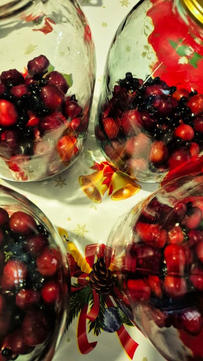 The image depicts a close-up, overhead view of four glass jars filled with red and black berries, possibly cranberries and blueberries, arranged on a festive tablecloth. The tablecloth features a repeating pattern of golden snowflakes and subtle Christmas-themed illustrations, including golden bells and red ribbons with pinecones and ornaments. The berries are plump and appear to be in a liquid, potentially being preserved or prepared for a holiday beverage or dish. The lighting creates highlights and shadows, emphasizing the reflective surfaces of the glass jars and the glossy texture of the berries. The scene appears to be set indoors, likely in a home environment, given the domestic nature of the objects and the Christmas decor. No people are visible in the image. The location is stated to be in the city of Troițcoe, Moldova.
