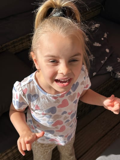 A young girl with blonde hair tied in a ponytail is looking up at the camera. She is wearing a t-shirt with a heart pattern and light-colored pants. She has a big smile on her face, showing her teeth. The image appears to be taken outdoors, with natural light casting shadows. The background consists of dark furniture and wooden planks, suggesting an outdoor seating area or deck. The specific location in Waldbröl, Germany is not discernible from the visual information provided.